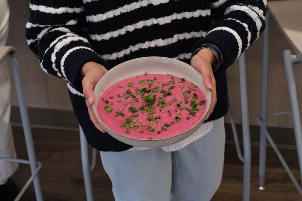 Second Harvest staff holding a bowl of vibrant beet and white bean hummus made from dry goods garnished with chopped herbs and pumpkin seeds. The individual is wearing a black-and-white striped sweater and light blue jeans