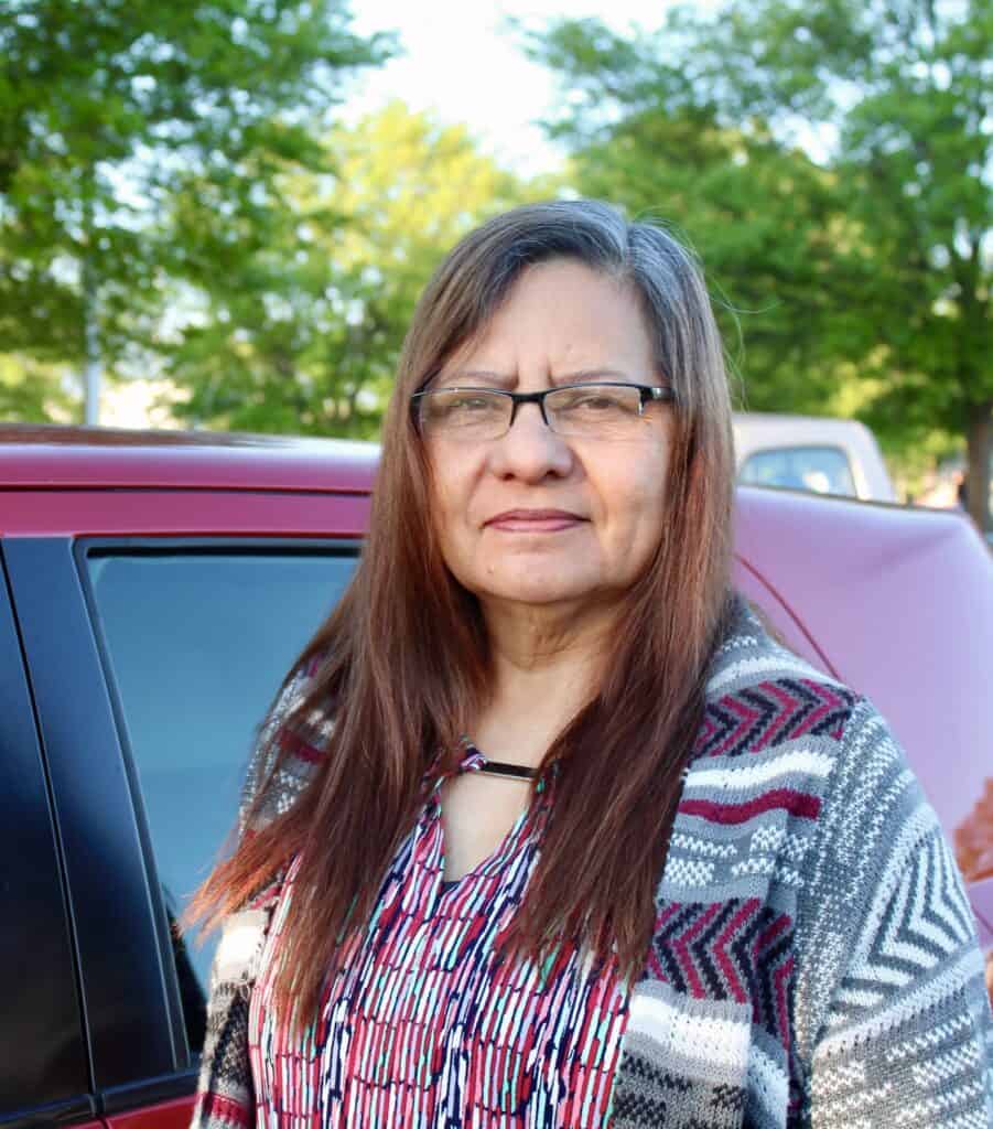 Juana, a woman facing the struggles of poverty, stands in front of a car at a food distribution with trees in the background on a sunny day.